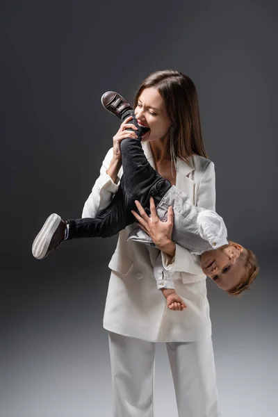 Збуджена жінка грає з маленьким сином і кусає ногу на сірому — стокове фото