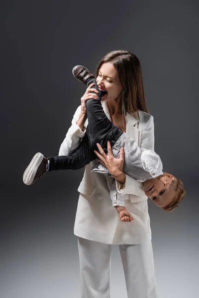 Excited woman playing with little son and biting his leg on grey — Photo de stock