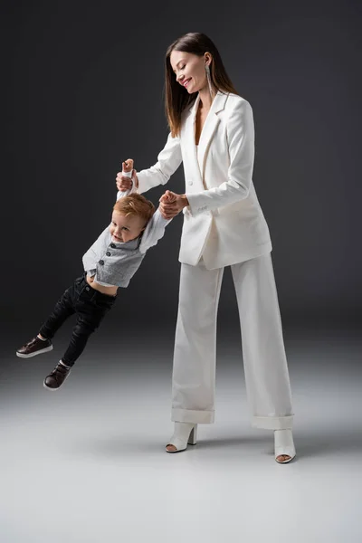 Full length view of cheerful woman holding hands of son while lifting him on grey — Stockfoto