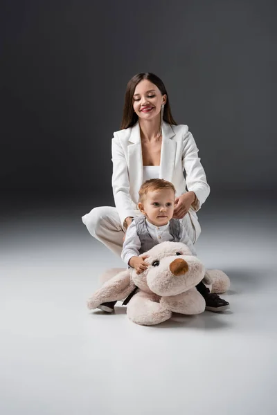 Baby boy sitting on toy dog near happy mom in white suit on grey — Photo de stock