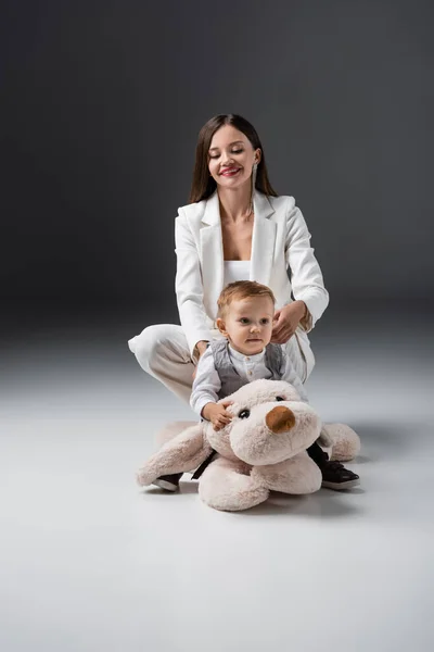 Baby boy sitting on toy dog near happy mom in white suit on grey — Stockfoto