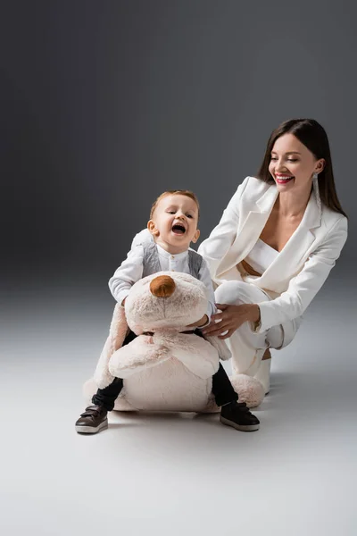 Aufgeregter Junge sitzt auf riesigem Spielzeughund neben glücklicher und stilvoller Mutter auf grau — Stockfoto