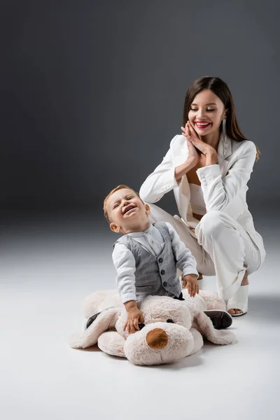 Fröhlicher Junge lacht auf riesigem Spielzeughund neben lächelnder Mutter auf grau — Stockfoto