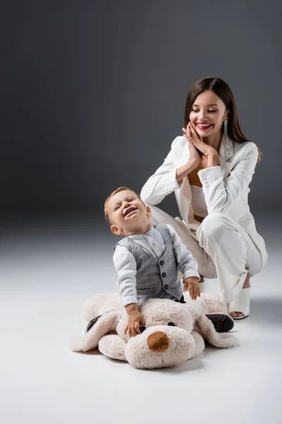 Alegre chico riendo en enorme juguete perro cerca sonriente madre en gris - foto de stock