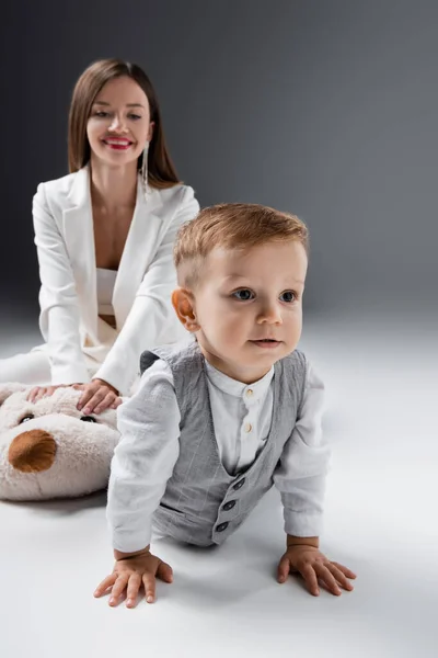 Little boy in stylish clothes crawling near mother smiling on blurred background on grey — Fotografia de Stock