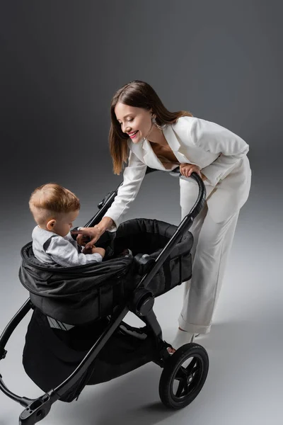 Happy woman touching toddler son sitting in black pram on grey — Stock Photo