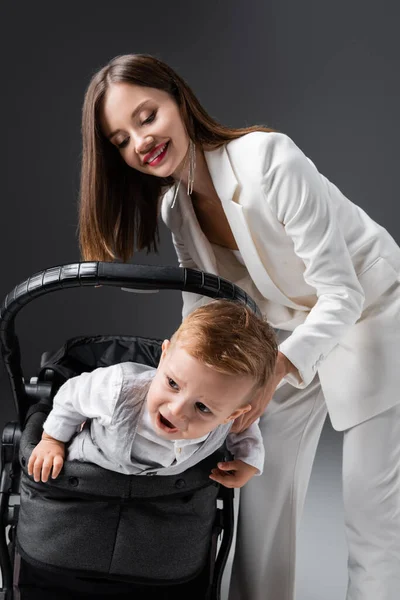 Trendfrau lächelt neben aufgeregtem Sohn im Kinderwagen auf grau — Stockfoto