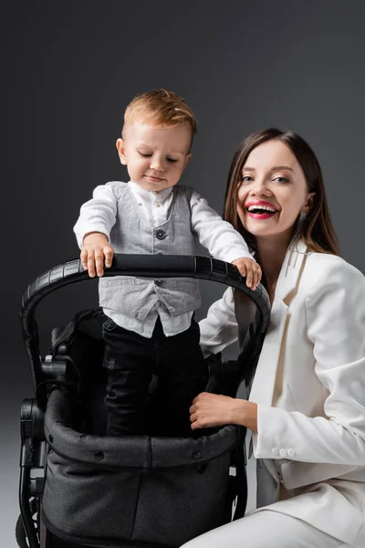 Joyful woman looking at camera near stylish son in baby carriage on grey — Stockfoto