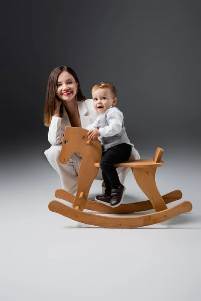 Amazed boy riding rocking horse near happy young mother on grey — Photo de stock