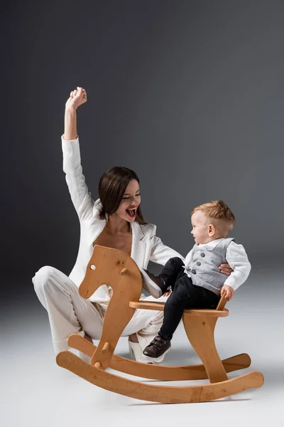 Excited mother showing triumph gesture near little son on rocking horse on grey — Stock Photo