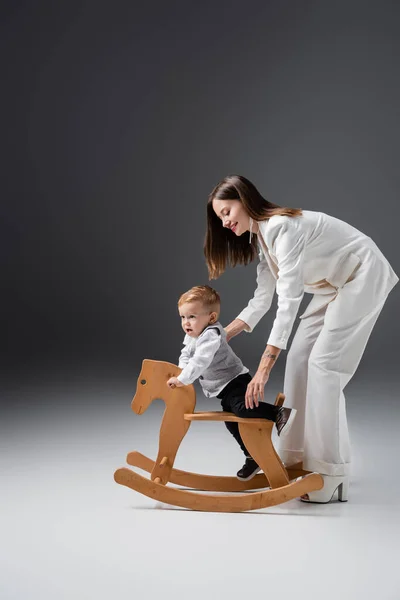 Vista de longitud completa de la mujer sonriente de apoyo bebé niño cerca de balanceo caballo en gris - foto de stock