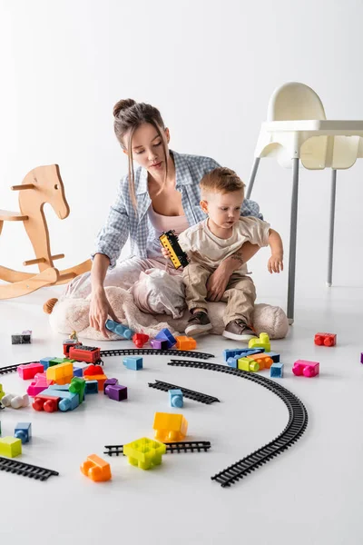 Jovem mulher segurando pequeno filho perto brinquedo ferroviário no branco — Fotografia de Stock