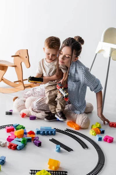 Маленький хлопчик з молодою матір'ю грає з іграшковою залізницею на білому — стокове фото