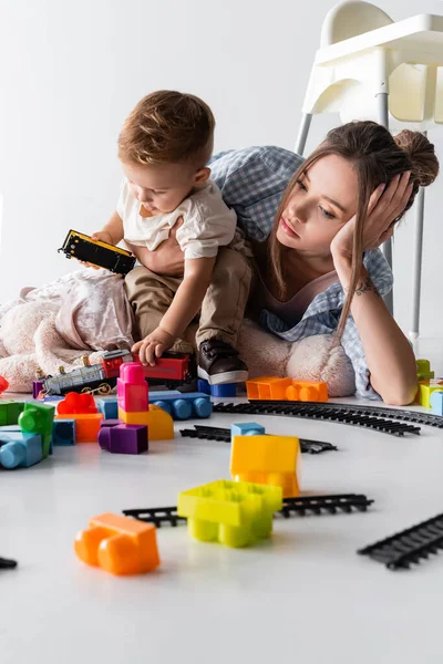 Müde Mutter liegt neben Kleinkind Sohn spielt mit Spielzeugeisenbahn auf weiß — Stockfoto