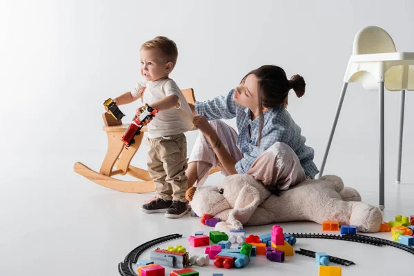 Unzufriedener Junge hält Spielzeugeisenbahn neben Mutter, die auf Grau sitzt — Stockfoto