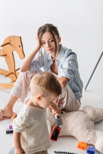 Müde Frau blickt auf Kleinkind-Sohn, der auf verschwommenem Vordergrund auf Weiß spielt — Stockfoto