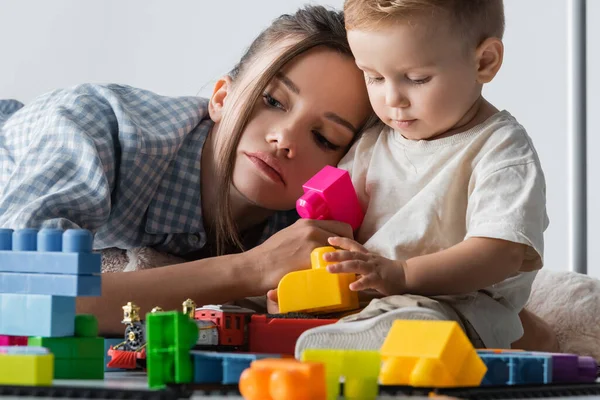 Kleiner Junge spielt mit Bauwürfeln nahe müder Mutter auf grau — Stockfoto