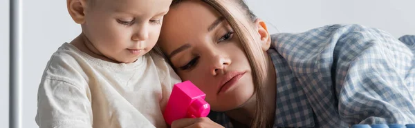 Baby boy holding construction cube near tired mom on grey, banner — Stock Photo