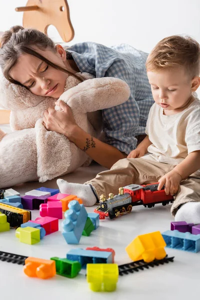 Donna accigliata che abbraccia giocattolo morbido vicino al figlio del bambino che gioca sul bianco — Foto stock