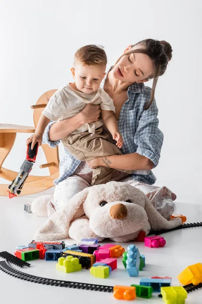 Frau umarmt Baby Junge, während sie auf großen Spielzeughund auf weiß sitzt — Stockfoto