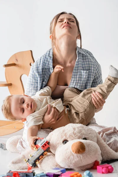 Femme épuisée tenant son fils près du chien jouet et cheval à bascule sur blanc — Photo de stock