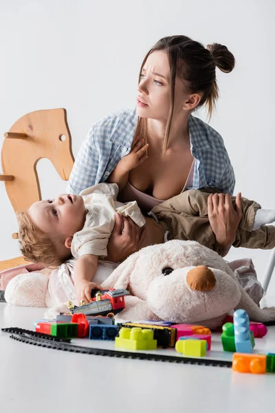 Femme fatiguée regardant loin tout en tenant son fils près de jouets sur blanc — Photo de stock