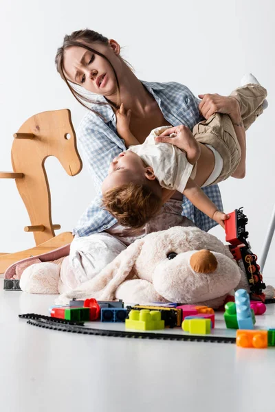 Femme fatiguée tenant fils tout en jouant avec lui près de jouets sur blanc — Photo de stock