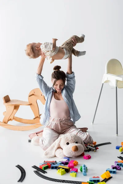 Mujer alegre sosteniendo pequeño hijo por encima de la cabeza mientras está sentado en juguete perro en blanco - foto de stock