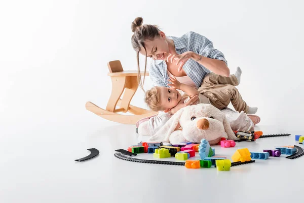 Mother showing scary gesture to little son while playing near toys on floor on white — стоковое фото