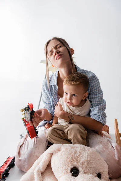 Femme fatiguée avec les yeux fermés embrassant fils et tenant train jouet sur blanc — Photo de stock