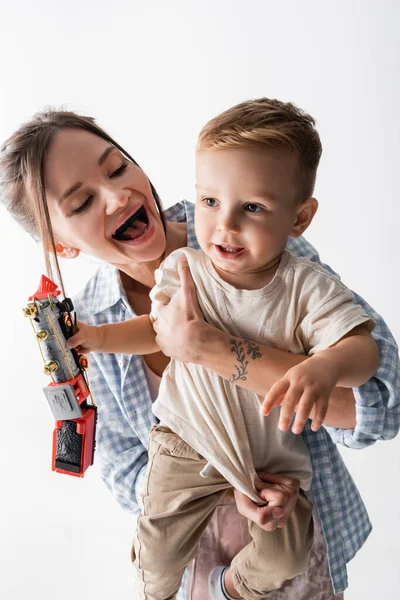 Cheerful woman embracing toddler son holding toy train isolated on white — Stockfoto