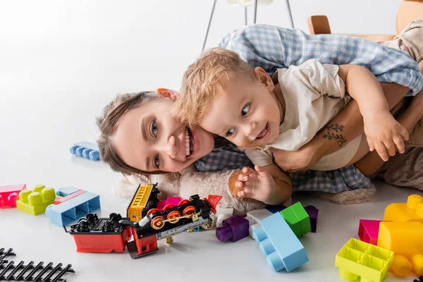 Femme gaie embrassant fils tout en étant couché près de jouets sur blanc — Photo de stock