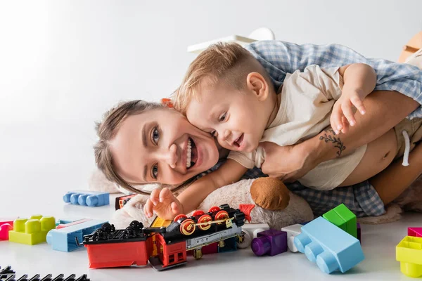 Cheerful woman looking at camera while hugging toddler son near toys on white — Photo de stock