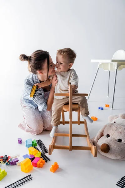 Junge reitet Schaukelpferd neben glücklicher Mutter und Spielzeug auf weißem Grund — Stockfoto