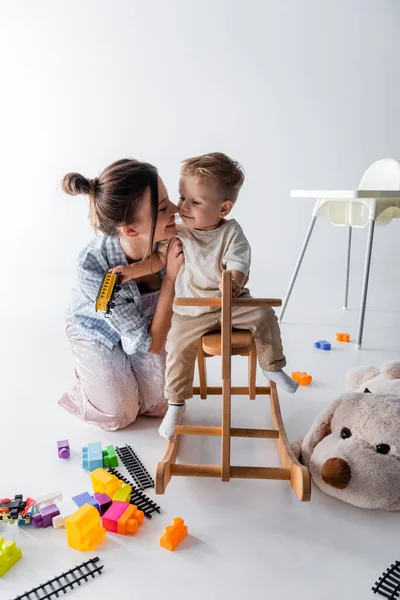 Bambino ragazzo cavalcando cavallo a dondolo vicino felice madre e giocattoli su bianco — Foto stock