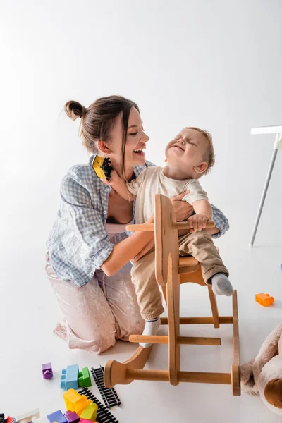 Glückliche Frau umarmt fröhlichen Sohn Reiten Schaukelpferd auf weiß — Stockfoto