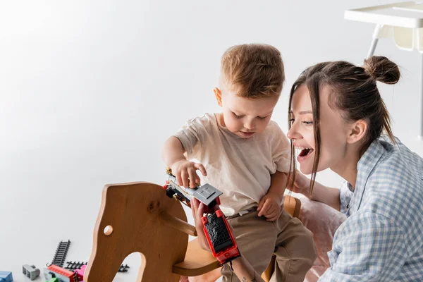 Pequeño niño sosteniendo juguete tren cerca asombrado madre en gris - foto de stock