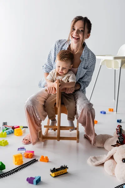 Excited woman looking at camera while riding rocking horse with son on white — Fotografia de Stock