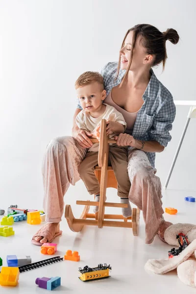 Junge und glückliche Frau reitet Schaukelpferd zusammen mit kleinem Sohn auf weiß — Stockfoto