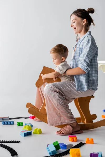 Side view of happy woman with son riding rocking horse on grey — Stock Photo