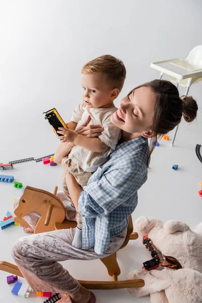 High angle view of joyful woman riding rocking horse together with son on white - foto de stock