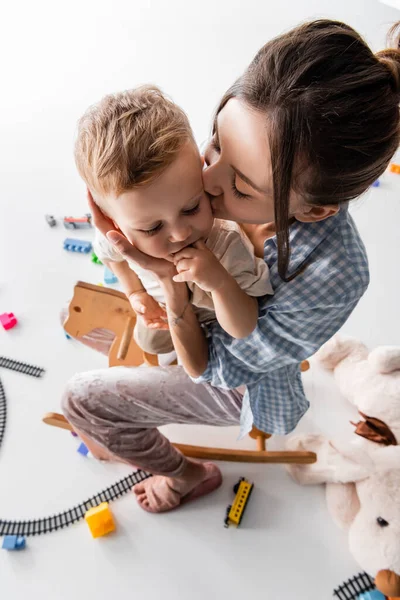 Вид зверху на жінку, яка цілує маленького сина біля іграшок на білому — стокове фото