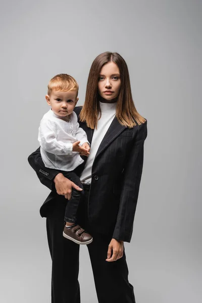 Femme d'affaires en costume tenant tout-petit enfant et regardant la caméra isolée sur gris — Photo de stock