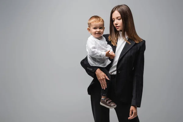 Giovane donna d'affari che tiene piccolo figlio sorridente alla fotocamera isolata su grigio — Foto stock