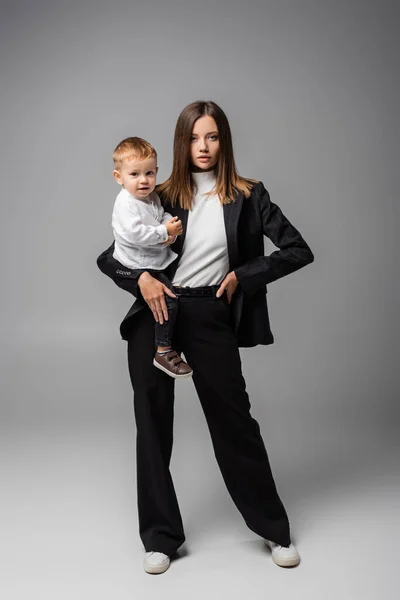 Businesswoman in black suit standing with toddler son on grey — стоковое фото