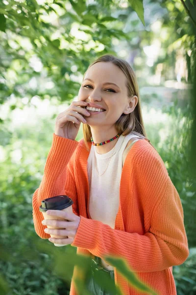 Joyful woman holding paper cup with coffee to go and touching lips in green park — Stock Photo