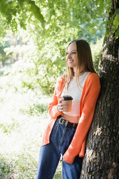 Joyful woman holding paper cup with coffee to go and standing near tree trunk in park — Fotografia de Stock