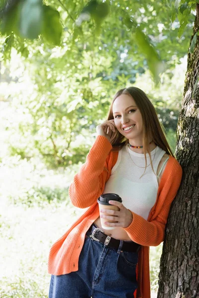 Mulher satisfeita segurando xícara de papel com café para ir e ficar perto de tronco de árvore — Fotografia de Stock