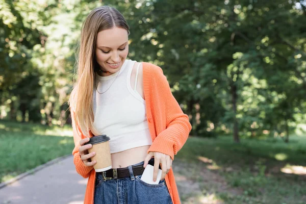 Pleased woman holding paper cup while taking smartphone from pocket in green park — Stock Photo