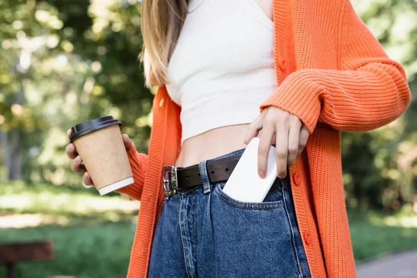 Vista recortada de la mujer sosteniendo la taza de papel mientras toma el teléfono inteligente de bolsillo - foto de stock